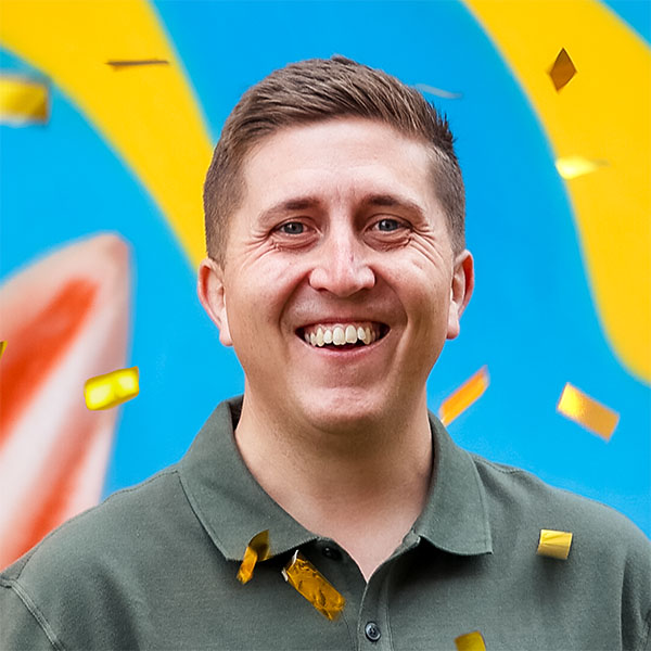 A smiling man in a green shirt stands against a vibrant blue background with yellow and orange abstract shapes, while golden confetti falls around him.