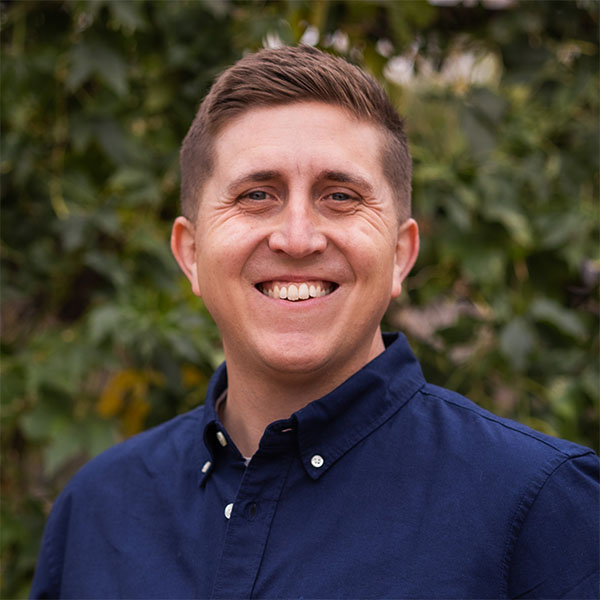 Smiling man wearing a dark blue button-up shirt stands outdoors with greenery in the background.