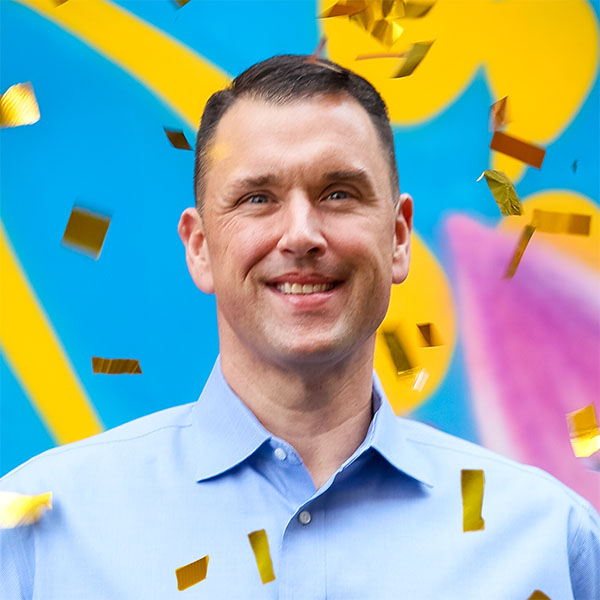 A smiling man in a light blue shirt stands against a vibrant background, with golden confetti falling around him.