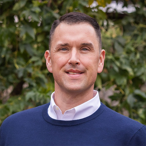 A man with short hair and a slight smile stands against a backdrop of green foliage, wearing a navy blue sweater and a white collared shirt.