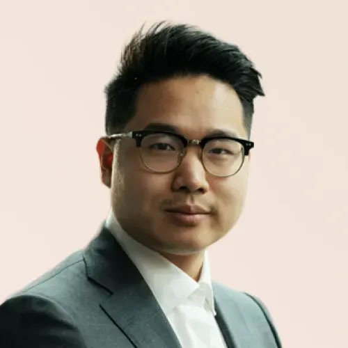 Portrait of a young man in a suit with glasses, smiling confidently against a soft pink background.