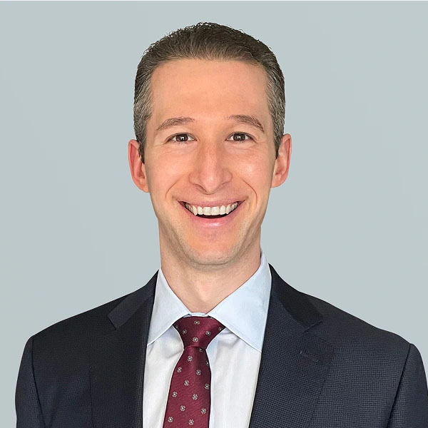 A man with short, styled hair smiles confidently while wearing a dark suit, a light blue shirt, and a maroon tie with a pattern. The background is a light gray.
