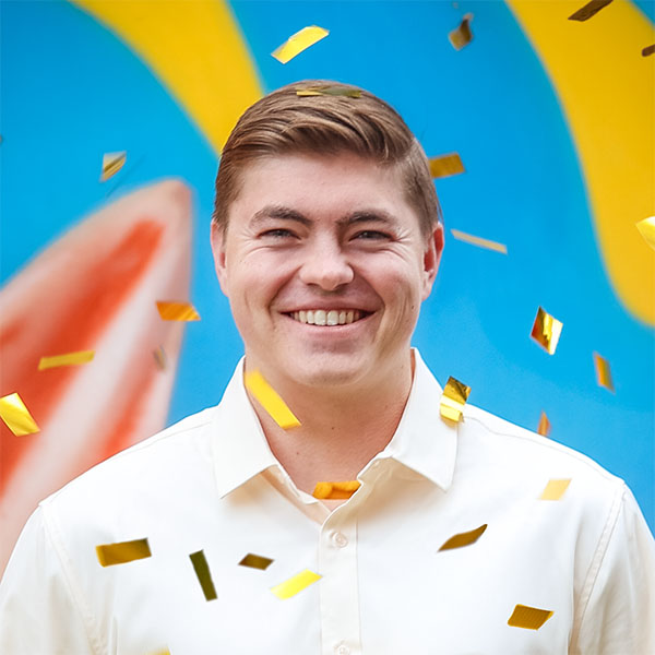 A smiling young man in a light-colored shirt stands in front of a vibrant blue background, surrounded by falling gold confetti.