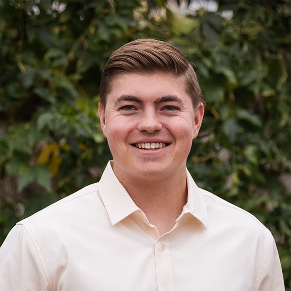 A young man with short brown hair is smiling while wearing a light-colored button-up shirt, standing in front of a green, leafy background.
