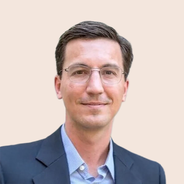 A man with short dark hair and glasses smiles confidently, wearing a dark blazer over a light blue shirt, set against a neutral background.