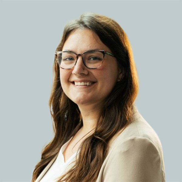 A woman with long, wavy brown hair and glasses smiles warmly, wearing a light-colored blazer against a pale gray background.