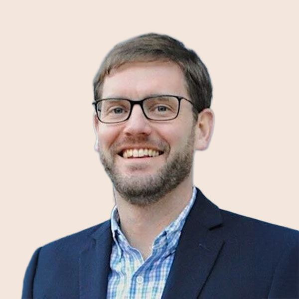 A smiling man with glasses and light brown hair, wearing a dark blazer over a checkered shirt, against a light pink background.
