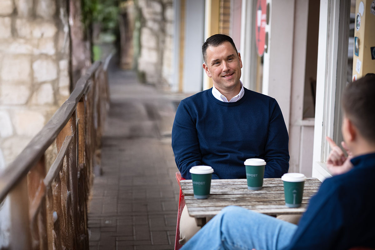 Two men are seated at a wooden table outdoors, with three coffee cups placed on the table. One man, wearing a blue sweater and white shirt, is smiling and looking at the other man, who is partially out of view and gesturing with his hand. The background features a stone wall and a walkway.