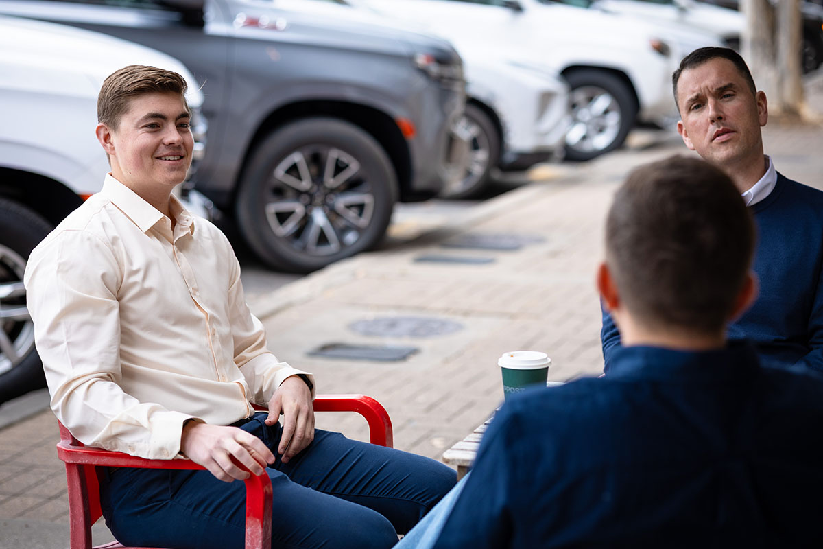 Aerial view of a cable-stayed bridge over a river during sunset, with vibrant colors in the sky and water, surrounded by lush greenery and urban landscapes in the background.