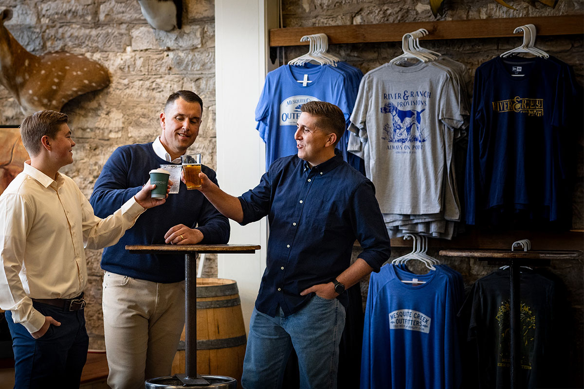 Three men are standing around a small table in a casual setting, smiling and toasting with drinks. One man holds a paper cup, another has a glass of beer, and the third man holds a glass with a light-colored drink. Behind them, a display of T-shirts hangs on the wall, and some animal mounts are visible.
