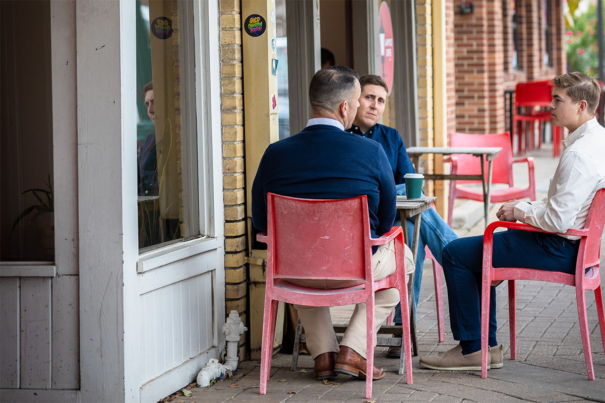 Three men sit at a small table outside a café, engaged in conversation. They are seated in red plastic chairs, with a green coffee cup placed on the table. A brick wall and large windows are visible in the background.