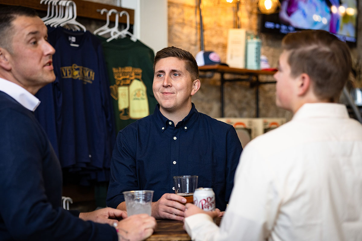 Three individuals engaged in conversation at a bar. One man in a navy shirt holds a drink, smiling, while the others, one in a white shirt and another in a dark sweater, are also attentive. Background shows clothing on hangers and a television.