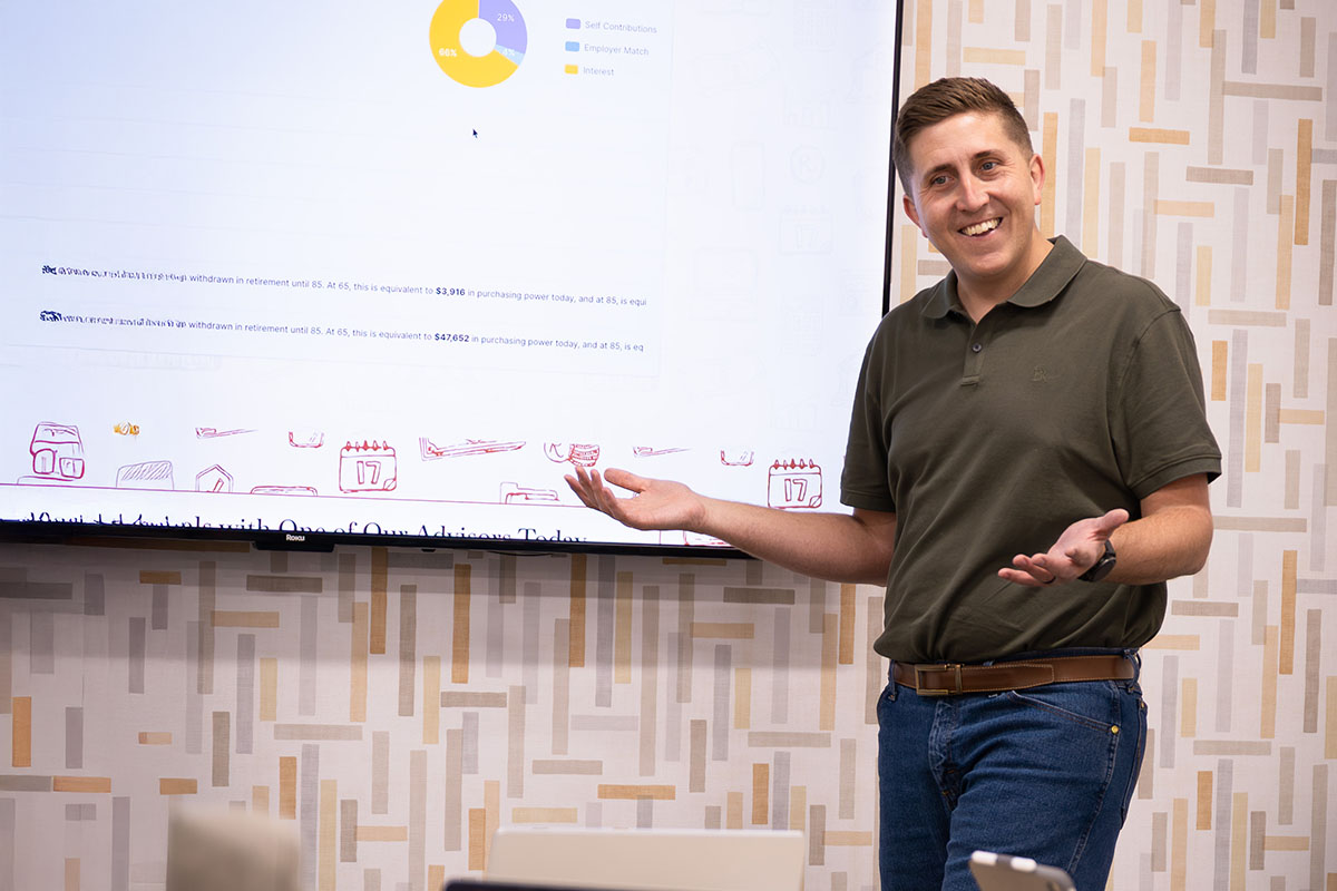 A man in a green polo shirt stands in front of a presentation screen, gesturing with his hands as he explains a financial chart. The screen displays a pie chart and text related to retirement withdrawals and purchasing power, with a patterned wall behind him.