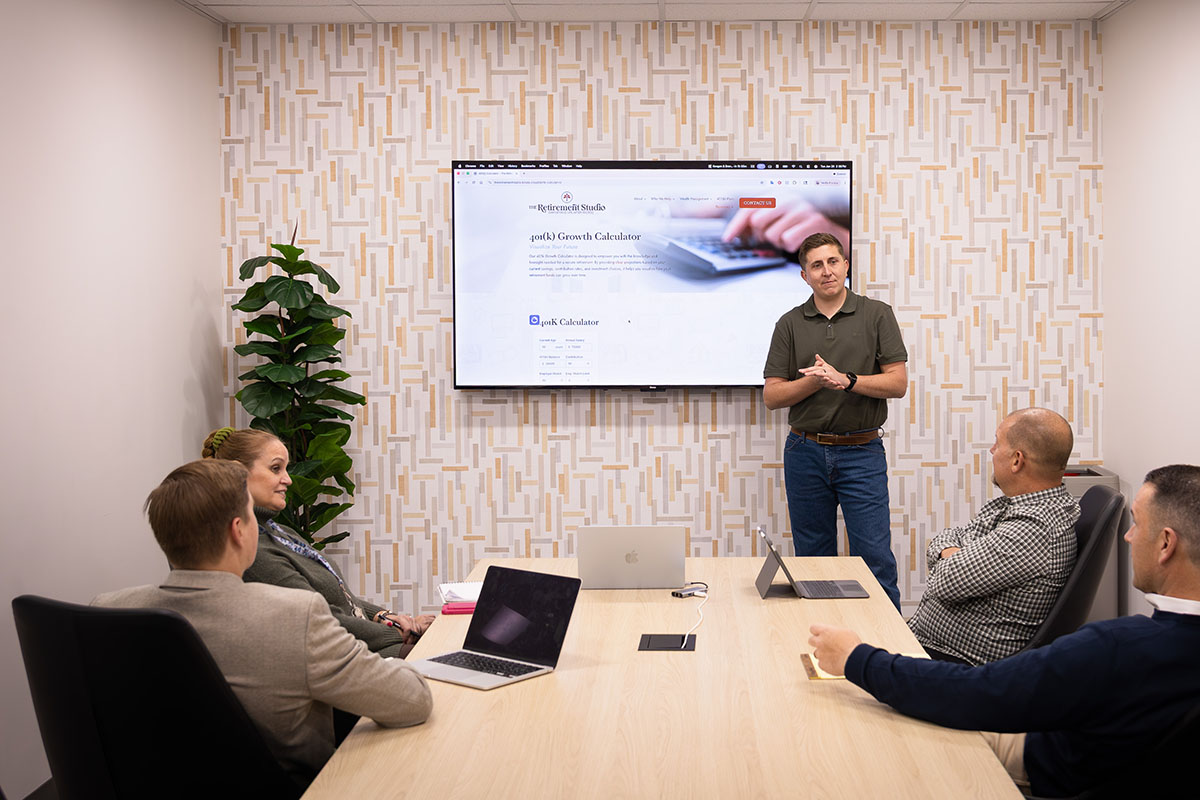 A man in a green shirt stands in front of a projector screen displaying a financial calculator website, while four others sit at a conference table with laptops, engaged in the presentation. A plant is visible in the background.
