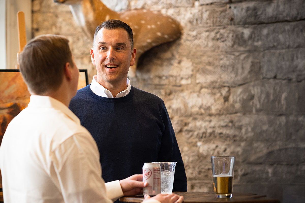 Two men are engaged in conversation at a bar, with one wearing a dark sweater and a collared shirt, appearing animated while speaking. The background features a stone wall and a mounted deer head, along with an easel displaying artwork. Drinks are on the table, including a can and glasses.