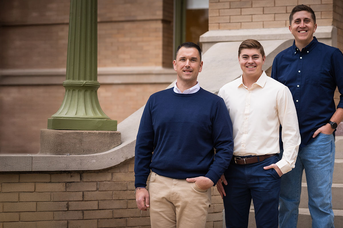 Three men stand together in front of a building, smiling. The man on the left wears a navy blue sweater and khaki pants, the middle man is dressed in a white button-up shirt and navy blue chinos, and the man on the right is in a dark blue shirt with light blue jeans. A green lamp post is visible beside them.