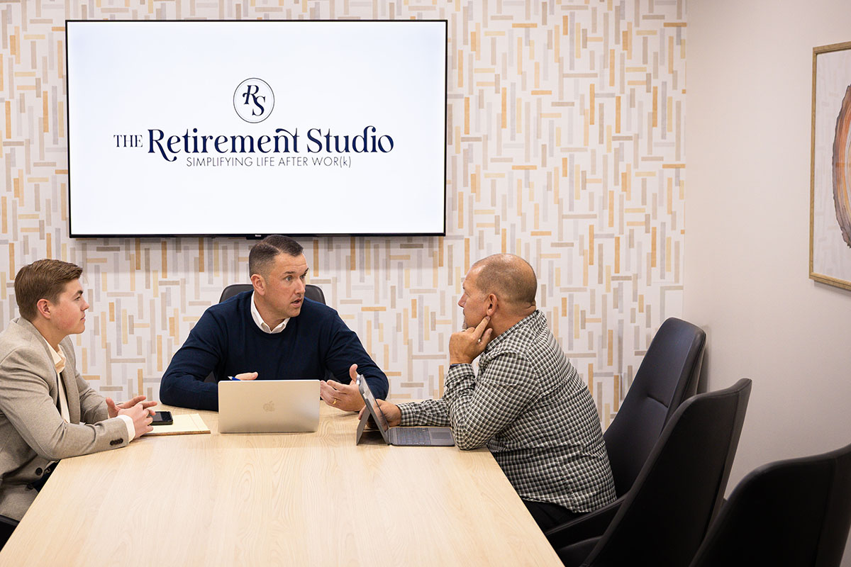 A meeting in a conference room featuring three men discussing around a wooden table, with a laptop and tablet in front of them. A large screen in the background displays "The Retirement Studio" logo, emphasizing their focus on retirement planning.